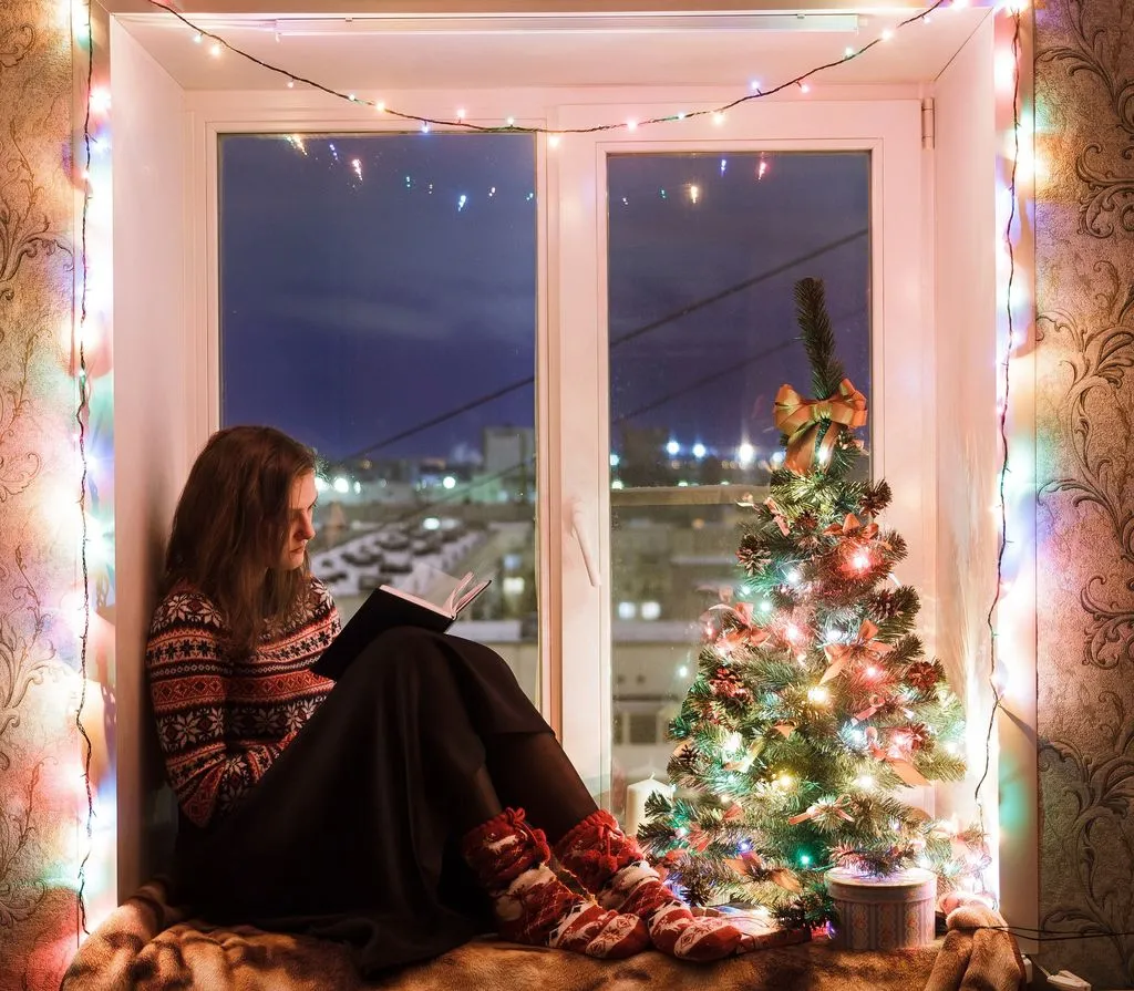 Young woman reading book sitting next to a lighted Christmas Tree near window with beautiful holiday decorations